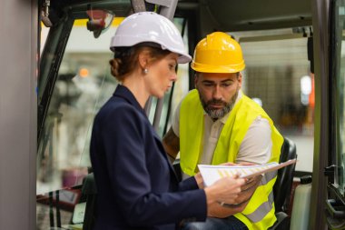 Erkek forklift sürücüsü modern endüstriyel fabrika müdürüyle konuşuyor. Depo görevlisi okuma emri, sipariş toplama. Depo çalışanı nakliyat, teslimat ve stokları kontrol etmek için ürün hazırlıyor.