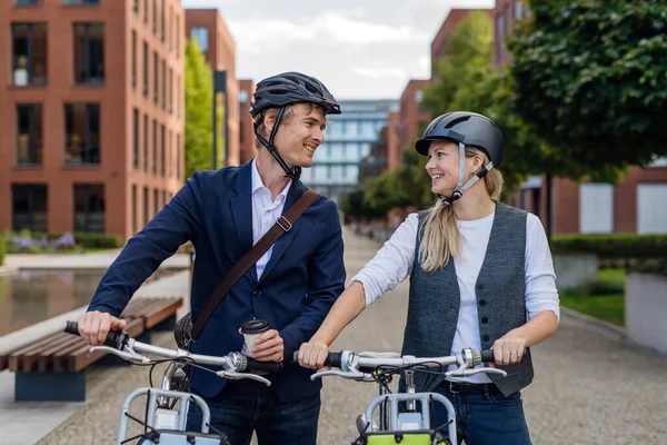 Spouses commuting through the city, talking and walking by bike on street. Middle-aged city commuters traveling from work by bike after a long workday. Husband and wife riding bikes in city.
