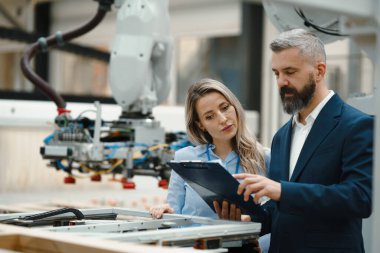Kadın mühendis ve erkek üretim müdürü modern endüstriyel fabrikada üretimden bahsediyorlar. Robot, robotik kollar ve otomasyon ile üretim tesisi.