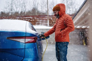 Soğuk karlı bir günde adam elektrikli arabayı şarj ediyor. Yakışıklı, olgun bir adamın soğuk havada şarj aletini şarj ettiği yer. Kış mevsiminde elektrikli araçların şarj edilmesi ve kullanılması.