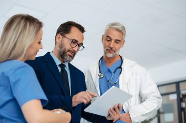 İlaç satış temsilcisi tıp binasındaki doktorlarla konuşuyor, onlara tablet üzerinde yeni ürünler sunuyor, iş konuşmaları yapıyor. Hastane müdürü salonda sağlık personeline danışmanlık yapıyor..