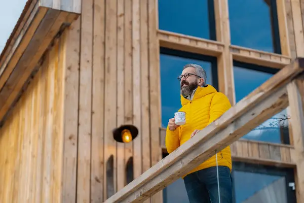 Mature man standing on cabins patio, drinking hot tea, coffee and enjoying beautiful winter day. Handsome man spending relaxing, stress-free winter weekend in cabin in mountains, enjoying alone time.