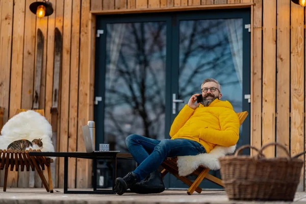 Man working from cozy cabin in mountains, sitting on terrace with laptop, enjoying cup of coffee, making call with client. Concept of remote work from beautiful, peaceful location. Hygge at work.