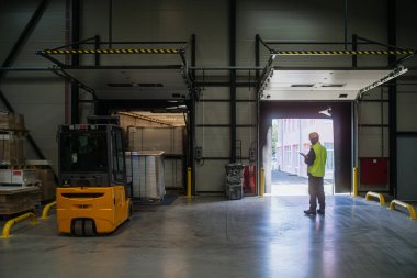 Depo alıcısı, Clark teslim edilen eşyaların depolanmasını denetliyor, tableti tutuyor, kargo detaylarına bakıyor. Forklift, içinde mallar olan paletler taşıyor.