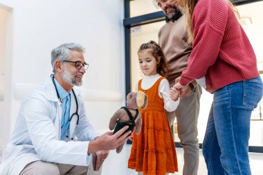 Pediyatrist kız oyuncağına bakıyor. Kız hasta, modern bir klinikte ailesiyle birlikte muayenede. Hastanede tıbbi muayene sırasında duygusal destek.