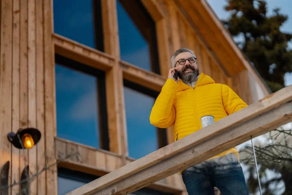 Mature man phone calling from patio, drinking hot tea, coffee and enjoying beautiful winter day. Handsome man spending relaxing, stress-free winter weekend in cabin in mountains, enjoying alone time.