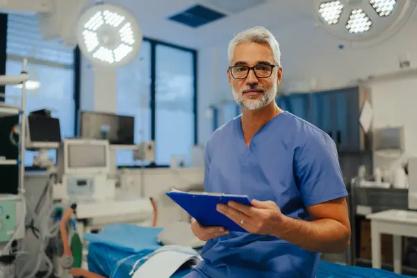 Kendine güvenen acil servis doktorunun portresi hastanede, acil serviste duruyor. Önlüklü yakışıklı doktor test sonuçlarıyla dolu bir pano tutuyor, modern özel klinikte kameraya bakıyor..