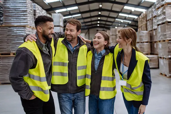 Full team of warehouse employees laughing in warehouse, holding each other by shoulders. Team of workers in reflective clothing in modern industrial factory, heavy industry, manufactrury. Group