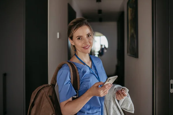 Female nurse, doctor getting ready for work, reading message on smartphone, leaving house in scrubs with backpack. Work-life balance for healthcare worker.