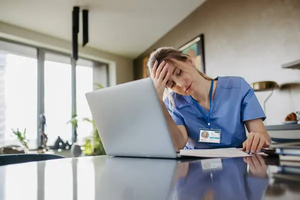 Nurse paying bills online, sitting in living room, working on notebook. Doctor in scrubs dealing with work issues from home, after workday, reading medical record, telemedicine consultation. Work-life