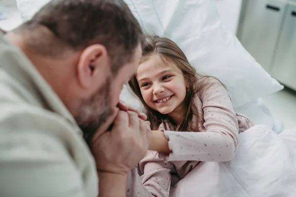 病院のベッドに横たわる娘のそばに座っている父親は プラッシュのおもちゃで遊んでいます 病院で病気の少女へのパパからの贈り物 — ストック写真