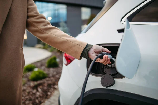 Close up of businessman charging electric car before going to office. An electric vehicle charging station in front of the office building. Charging at work, workplace.