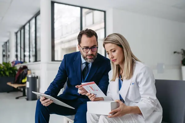 Portrait of pharmaceutical sales representative talking with doctor in medical building. Ambitious male sales representative in suit presenting new medication on tablet.