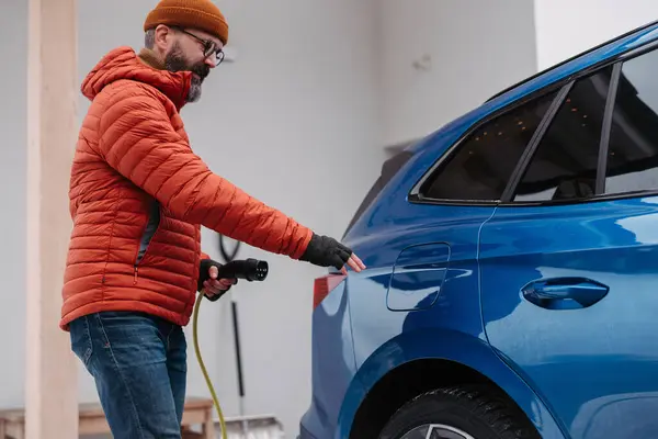 Man charging electric car during cold snowy day. Side view of hansome mature man putting charger in charging port during cold weather. Charging and driving electric vehicles during winter season.