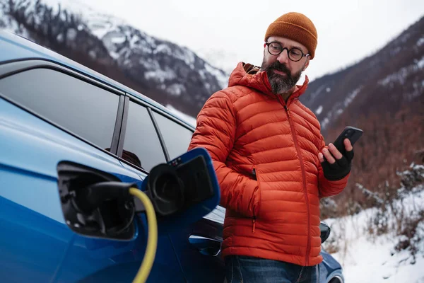 Mature man charging electric car during cold snowy day, using electric vehicle charging app, checking energy consumption, battery life on smart phone. Charging and driving electric vehicles during