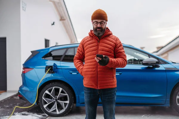 Mature man charging electric car during cold snowy day, using electric vehicle charging app, checking energy consumption, battery life on smart phone. Charging and driving electric vehicles during