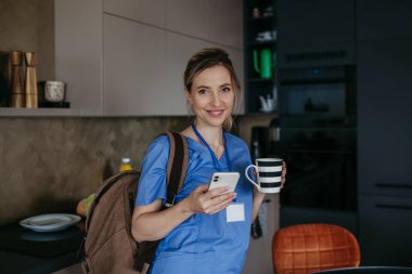 Kadın hemşire, doktor işe gitmek için hazırlanıyor, akıllı telefondan mesaj okuyor, kahve içiyor, ameliyat önlüğüyle evden ayrılıyor. Sağlık çalışanı için iş-yaşam dengesi.