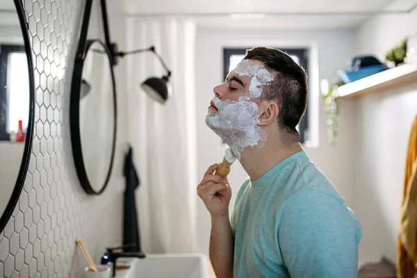 ダウン症候群の若者は 剃る方法を学び 剃る泡を塗り 顔全体にクリームをかけ 鏡を見る — ストック写真