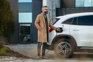 İşadamı ofise gitmeden önce elektrikli arabayı şarj ediyor. Ofis binasının önündeki elektrikli araç şarj istasyonu. İş yerinde, iş yerinde ücret..