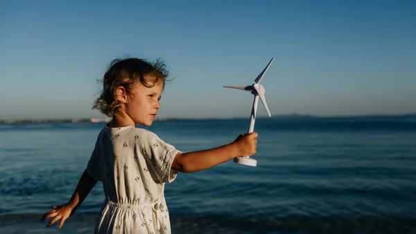 一个金发小女孩站在海滩上 手里拿着风力涡轮机模型 还有海风旋翼 — 图库照片