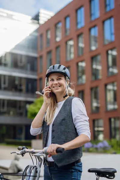 Beautiful middle-aged woman commuting through the city by bike, holding smartphone, phone calling. Female city commuter traveling from work by bike after a long workday.