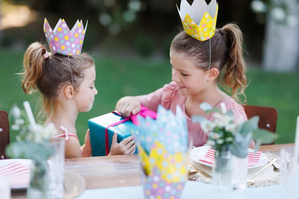 Little friends at a birthday party outdoors at the garden. Cute girls with paper crowns opening birthday gift. Birthday garden party for children.