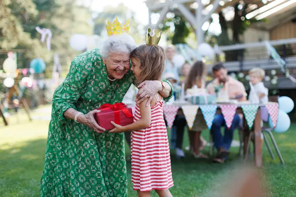 Garden birthday party for senior lady. Beautiful senior birthday woman receiving gift from granddaughter, hugging.
