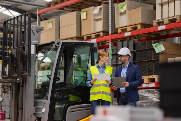 Female forklift driver talking with warehouse manager in suit, order picking. Warehouse worker preparing products for shipmennt, delivery, checking stock in warehouse.