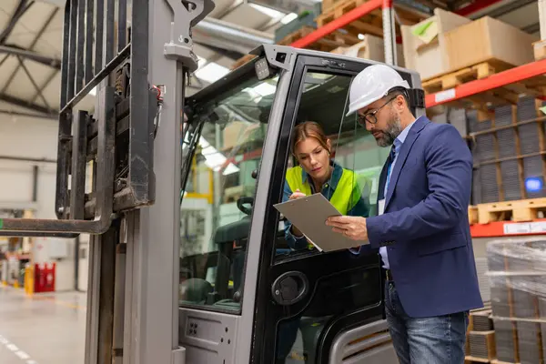 Female forklift driver talking with warehouse manager in suit, order picking. Warehouse worker preparing products for shipmennt, delivery, checking stock in warehouse.