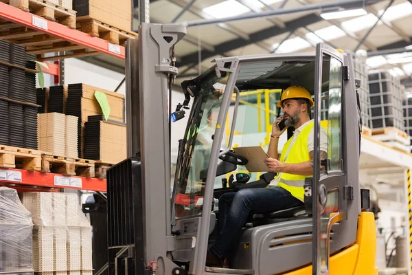 Warehouse forklift driver controlling shipping order, holding smartphone and clipboard. Warehouse worker preparing products for shipmennt, delivery, checking stock in warehouse, order picking.
