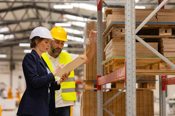 Female manager talking with foreman. Warehouse, production worker preparing products for shipment. Woman quality controller checking quality of products, talking with technician, holding clipboard