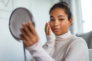 Güzel kıvrımlı bir kadının portresi, evdeki küçük yuvarlak makyaj aynasına bakıyor. Cilt rutini.