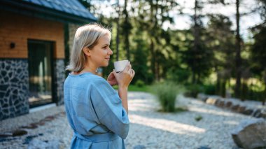 A happy young woman standing in garden near cottege and enjoying cup of morning coffee on summer vacation in mountains.