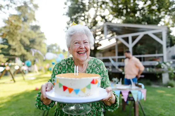 Beautiful senior birthday woman with a paper crown holding birthday cake. Garden birthday party for senior lady.