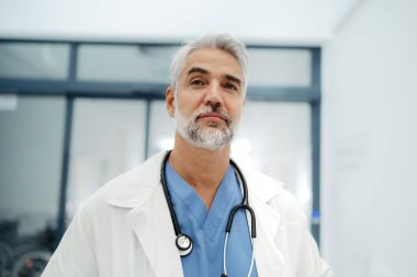 Hastane koridorunda duran kendinden emin olgun bir doktorun portresi. Beyaz önlüklü, önlüklü, steteskop takan yakışıklı bir doktor modern özel klinikte duruyor.