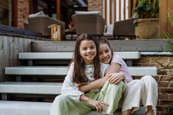 Two sisters sitting on front porch stairs in front of house, embracing. Sisterly love and siblings relationship concept.