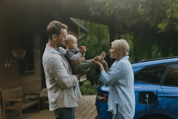 Young family with little baby standing by their electric car. Electric vehicle with charger in charging port.