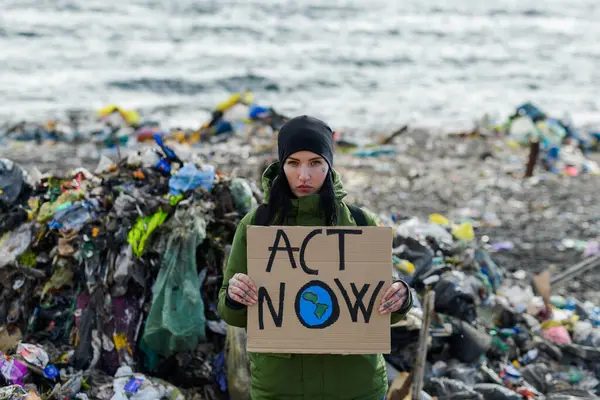 Woman activist holding placard, protest sign, standing on landfill, large pile of waste on sea beach, shoreline, environmental concept and eco activism.