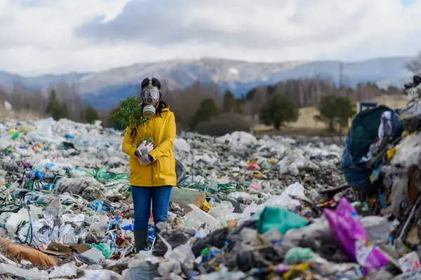 Female activist with gas mask holding potted plant standing on landfill, large pile of waste, environmental concept and eco activism.