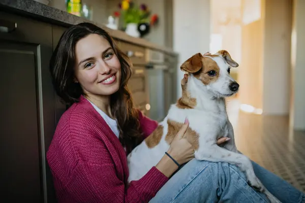 Young woman living alone in apartment. Beautiful single woman sitting on kitchen floor, petting her dog, enjoying weekend. Looking at camera, smiling.