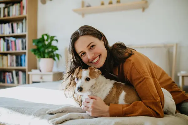 Young woman living alone in apartment. Beautiful single woman lying on bed, petting her dog, enjoying weekend. Looking at camera, smiling.