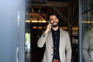 Takım elbiseli olgun iş adamının portresi modern ofiste kapalı bir yerde duruyor, telefonla konuşuyor..