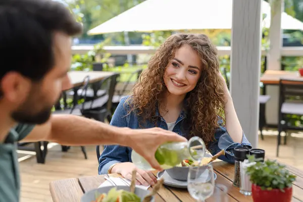 Young couple at date in restaurant, sitting on restaurant terrace. Boyfriend pouring water in girlfriends glass, taking care of her. Lunch or brunch outdoors, outdoor seating for dining.