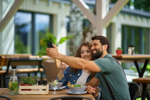 Young couple at date in restaurant, taking selfie, sitting on restaurant terrace. Boyfriend and girlfriend enjoying springtime, having lunch or brunch outdoors, outdoor seating for dining.