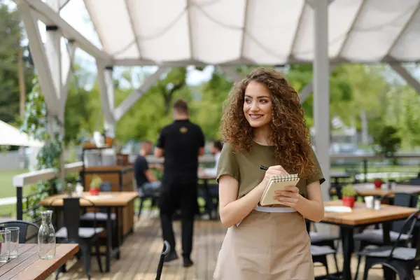 Portrait of a beautiful waitress writing down an order in paper notebook with pen. Female server standing on the restaurant terrace in an apron.