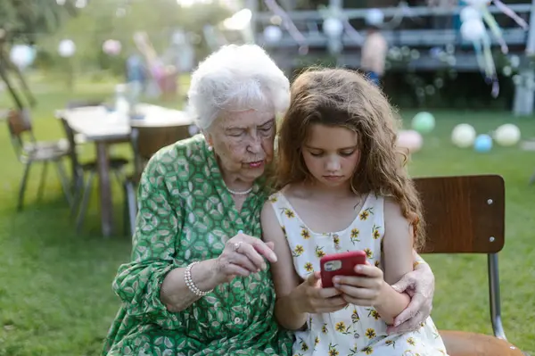 ガーデンパーティーで高齢のおばあちゃんにスマートフォンで何かを見せている少女 祖父母と孫の間の愛と親密さ — ストック写真