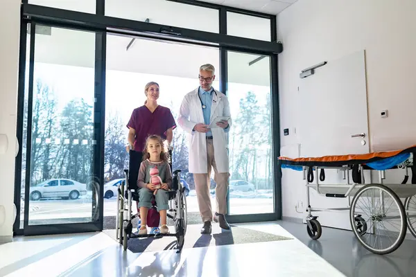小児科医は 車椅子で小さい患者と一緒に歩いています 車椅子を押す看護師さん 病院で医者と看護師を信頼するかわいい幼稚園の少女 子供の医療と感情の概念 — ストック写真