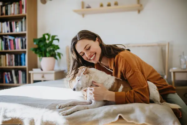 Young woman living alone in apartment. Beautiful single woman lying on bed, petting her dog, enjoying weekend, smiling.