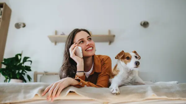 Young woman living alone in apartment, enjoying weekend. Beautiful single woman lying on bed, petting her dog, and making phone call.