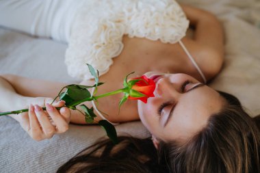 Yatakta uzanmış şehvetli bir kadın güzel kırmızı bir gül kokar. Sevgililer Günü kavramı, aşk ve romantik ilişki.